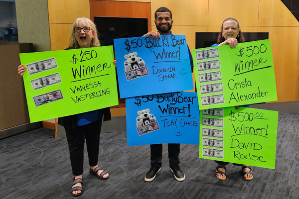 Three members of Ideal Staff holding signs for winners of events, $250 Winner Vanessa Vasterling, $50 Bucky Bear Winner Donovin Sheak, $50 Bucky Bear Winner Toby Chang, $500 Winner Crista Alexander and $500 winner David Rouse